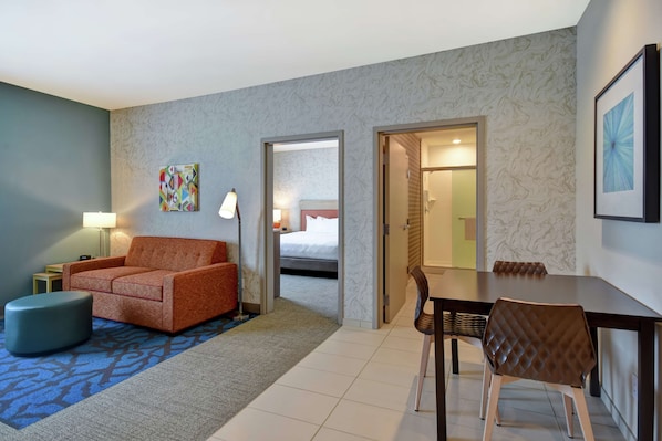 Habitación de hotel con sofá, reposapiés, mesa de comedor, sillas, cama y baño visible a través de una puerta abierta.
