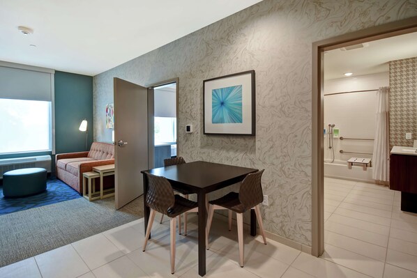 Una habitación de hotel moderna con zona de comedor, sofá y baño visible a través de una puerta abierta.