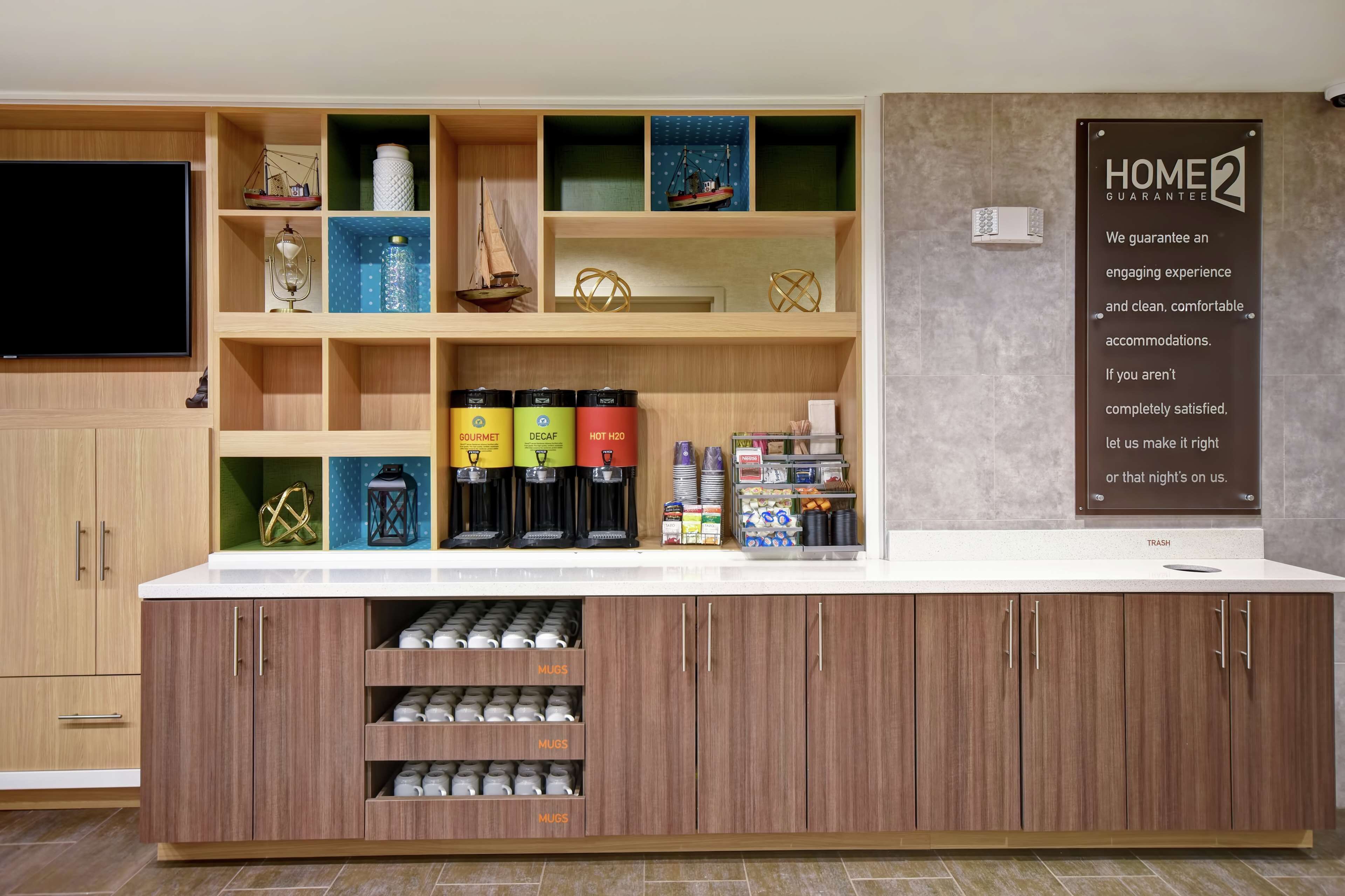 A hotel reception area with a counter, coffee machines, and a 'Home 2 Guarantee' sign.
