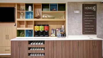 A hotel reception area with a counter, coffee machines, and a 'Home 2 Guarantee' sign.