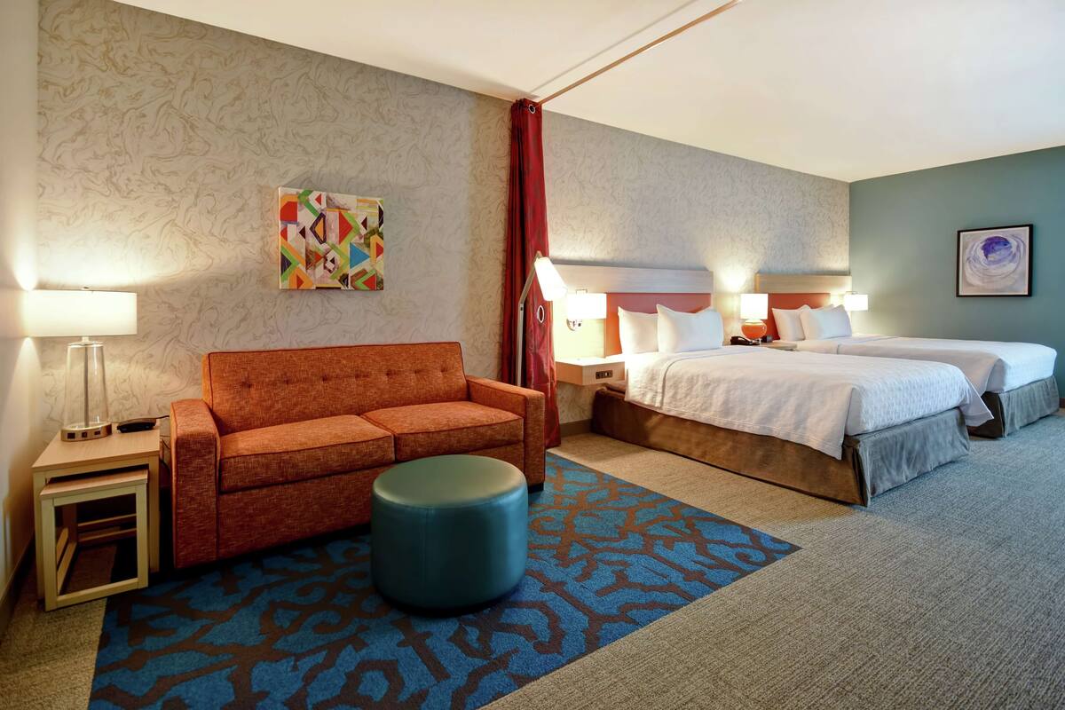 Ein Hotelzimmer mit einer Couch, einem Hocker, zwei Betten, einem Nachttisch, einer Lampe und einem Wandbild.