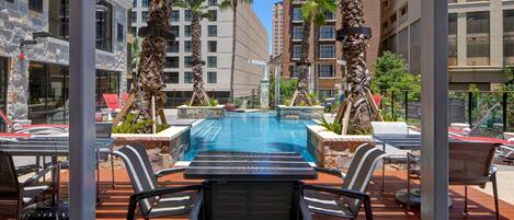 A pool area with lounge chairs and tables, surrounded by palm trees and a view of tall buildings.