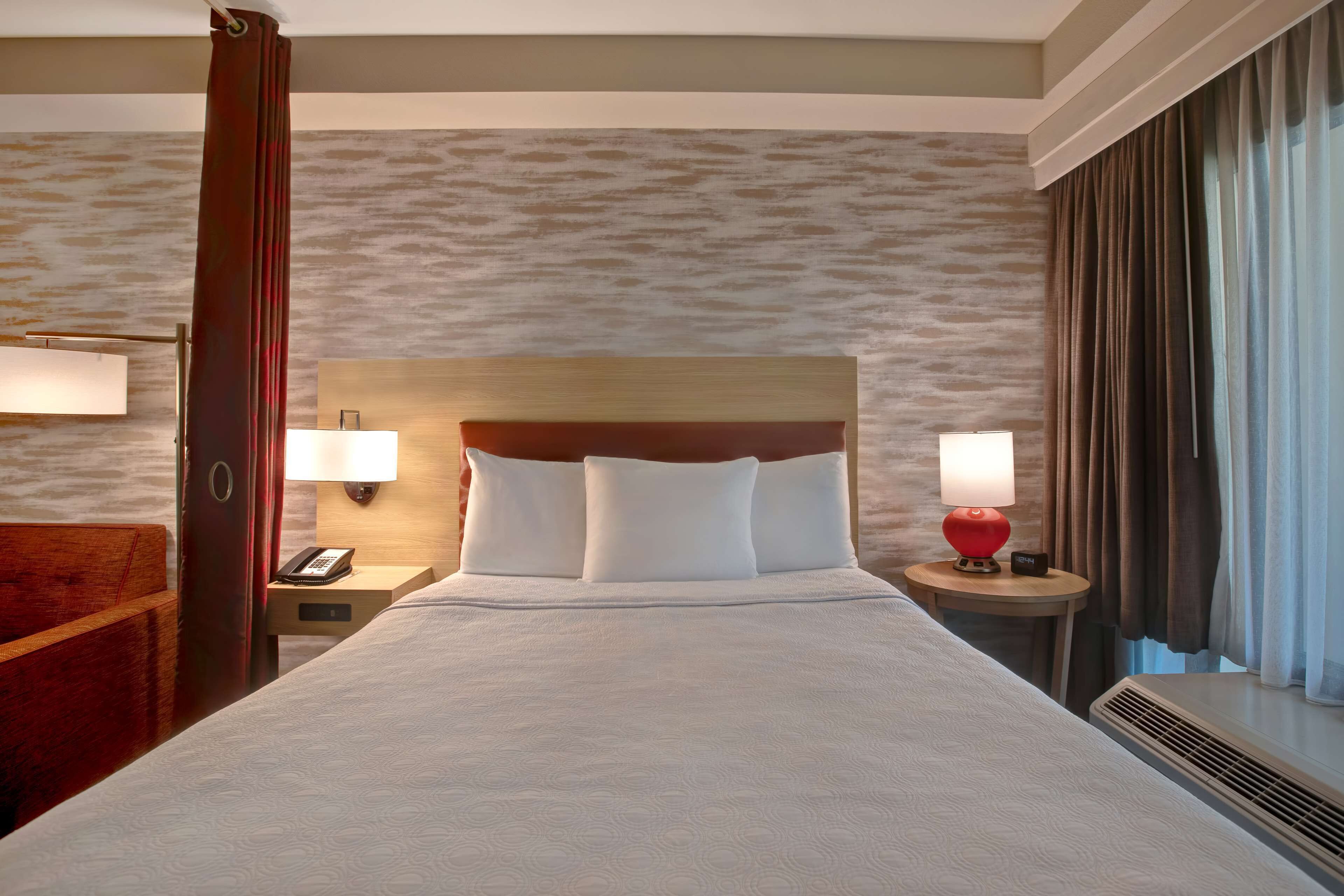 Quarto de hotel com uma cama grande, duas mesas de cabeceira, um painel de madeira na cabeceira, um abajur vermelho e uma janela com cortinas.