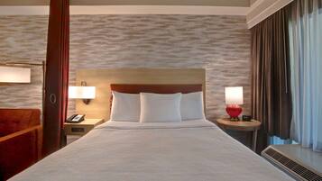 Quarto de hotel com uma cama grande, duas mesas de cabeceira, um painel de madeira na cabeceira, um abajur vermelho e uma janela com cortinas.