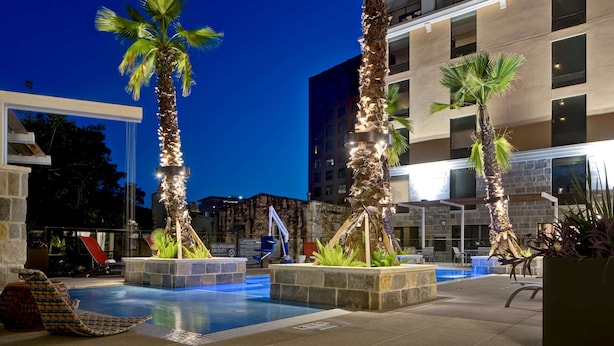 A modern hotel pool area with palm trees, lounge chairs, and a stone wall. - Home2 Suites by Hilton San Antonio Riverwalk, TX (San Antonio)