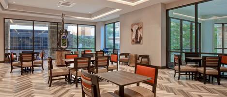 A spacious dining area with wooden flooring, multiple tables and chairs, large windows, and a hanging decorative piece.