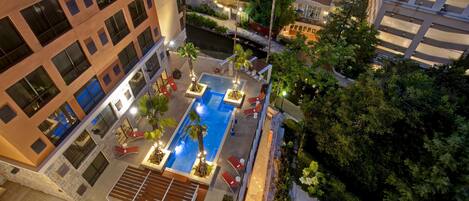 A rooftop pool area with a rectangular pool, sun loungers, and palm trees.