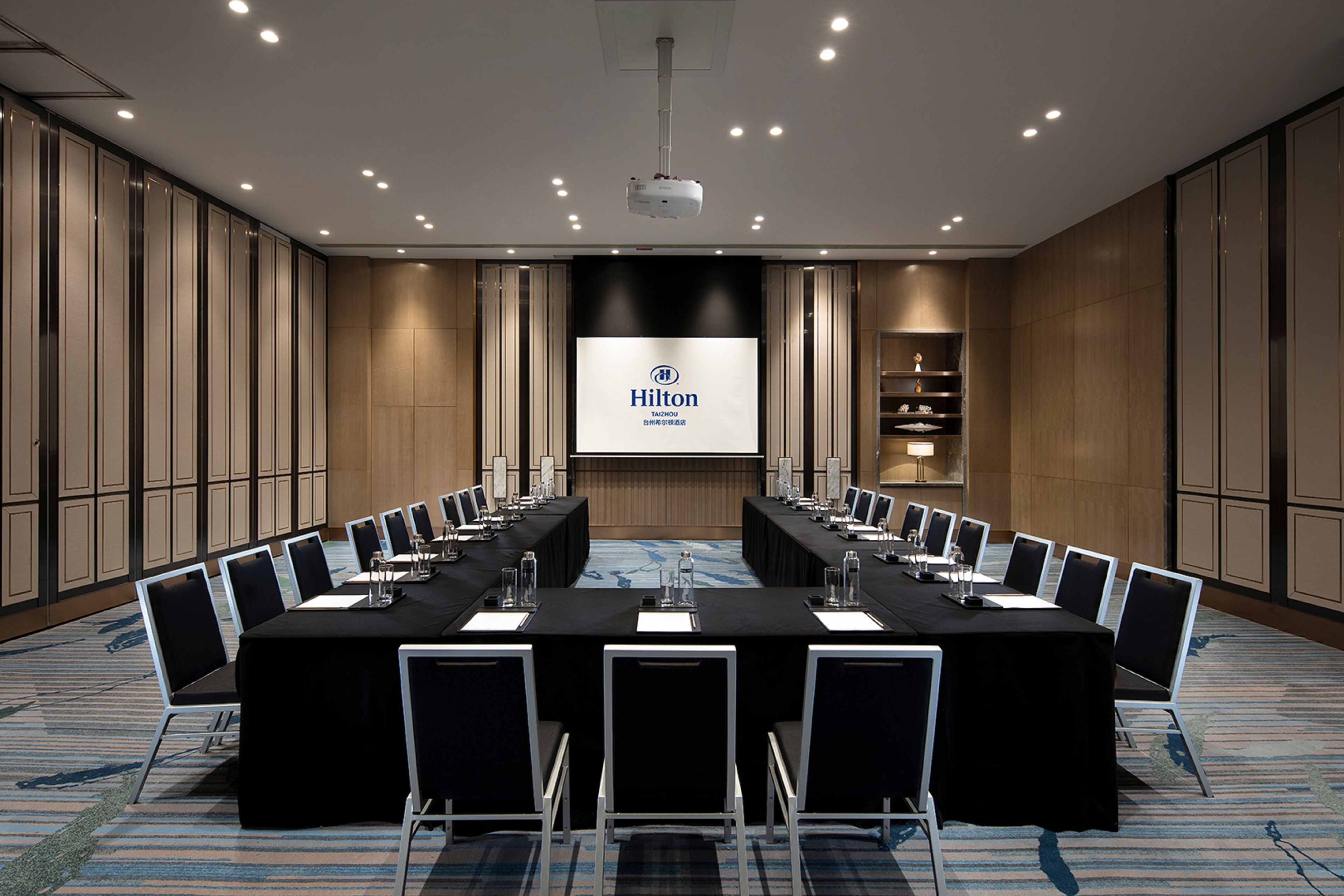 A conference room with a long table, chairs, a projector screen, and a presentation screen displaying the Hilton logo.