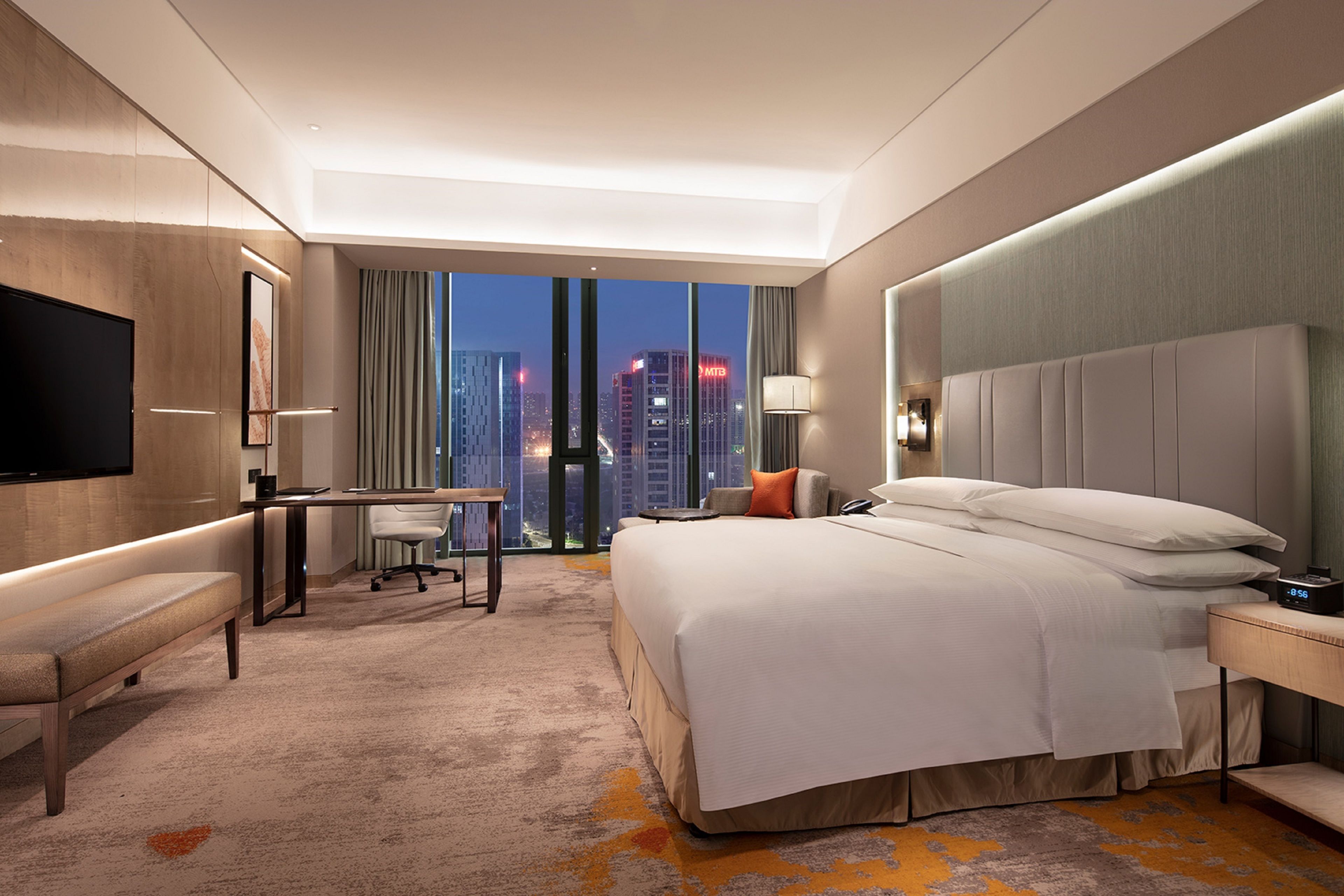 Ein modernes Hotelzimmer mit einem großen Bett, einem Schreibtisch, einem Sessel, einem Fernseher und Blick auf die Stadt.