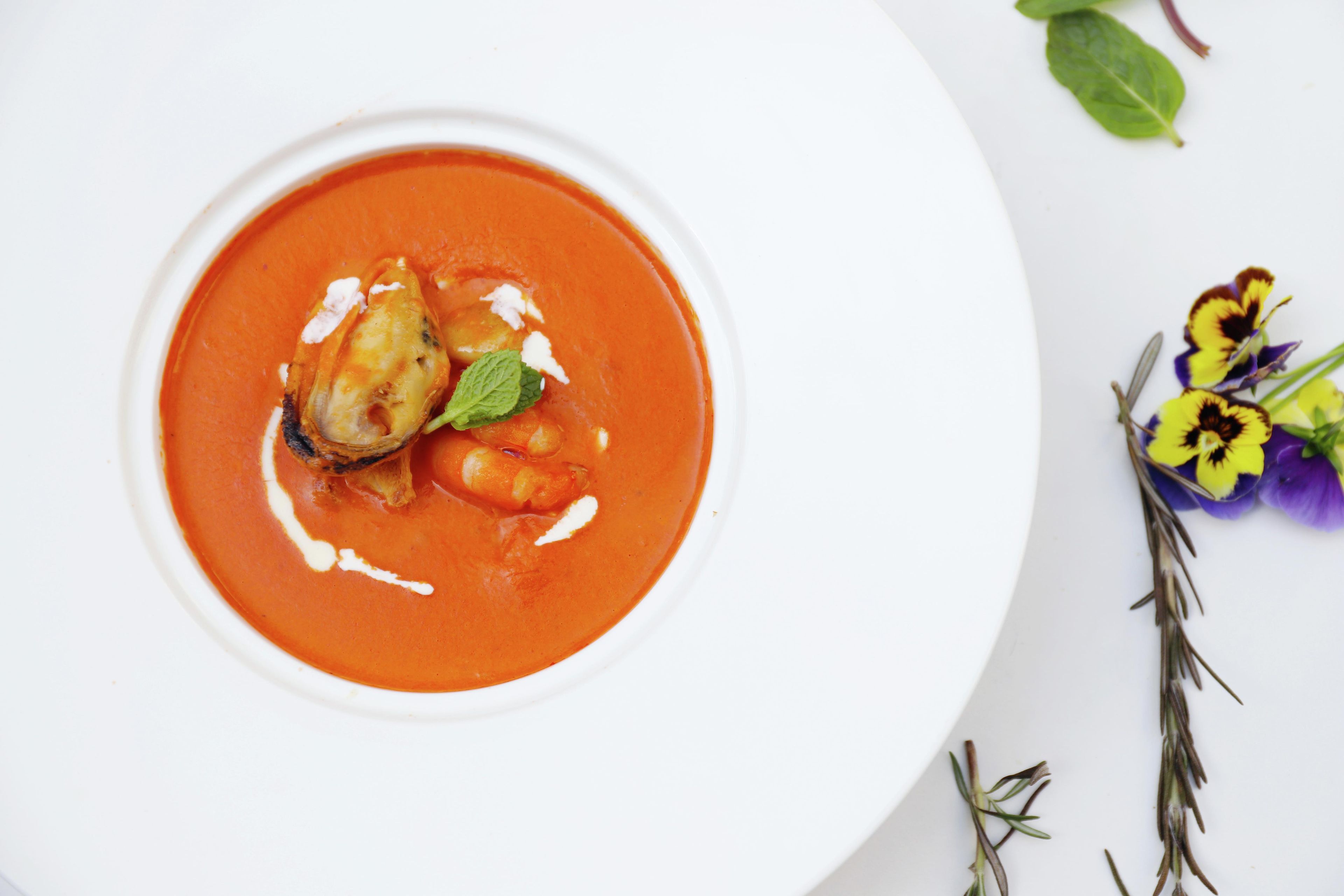 Un tazón de sopa con guarnición, acompañado de una ramita de romero y un pequeño ramo de flores sobre un plato blanco.