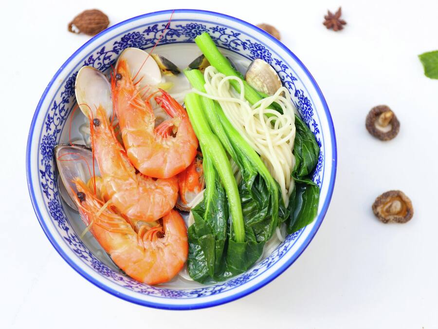 A bowl of soup with shrimp, noodles, and greens.