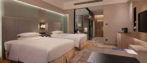 A modern hotel room with two beds, a desk, and a bathroom visible through a glass door.