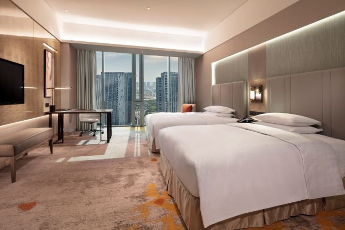 A hotel room with two beds, a desk, a TV, and a large window with a city view.