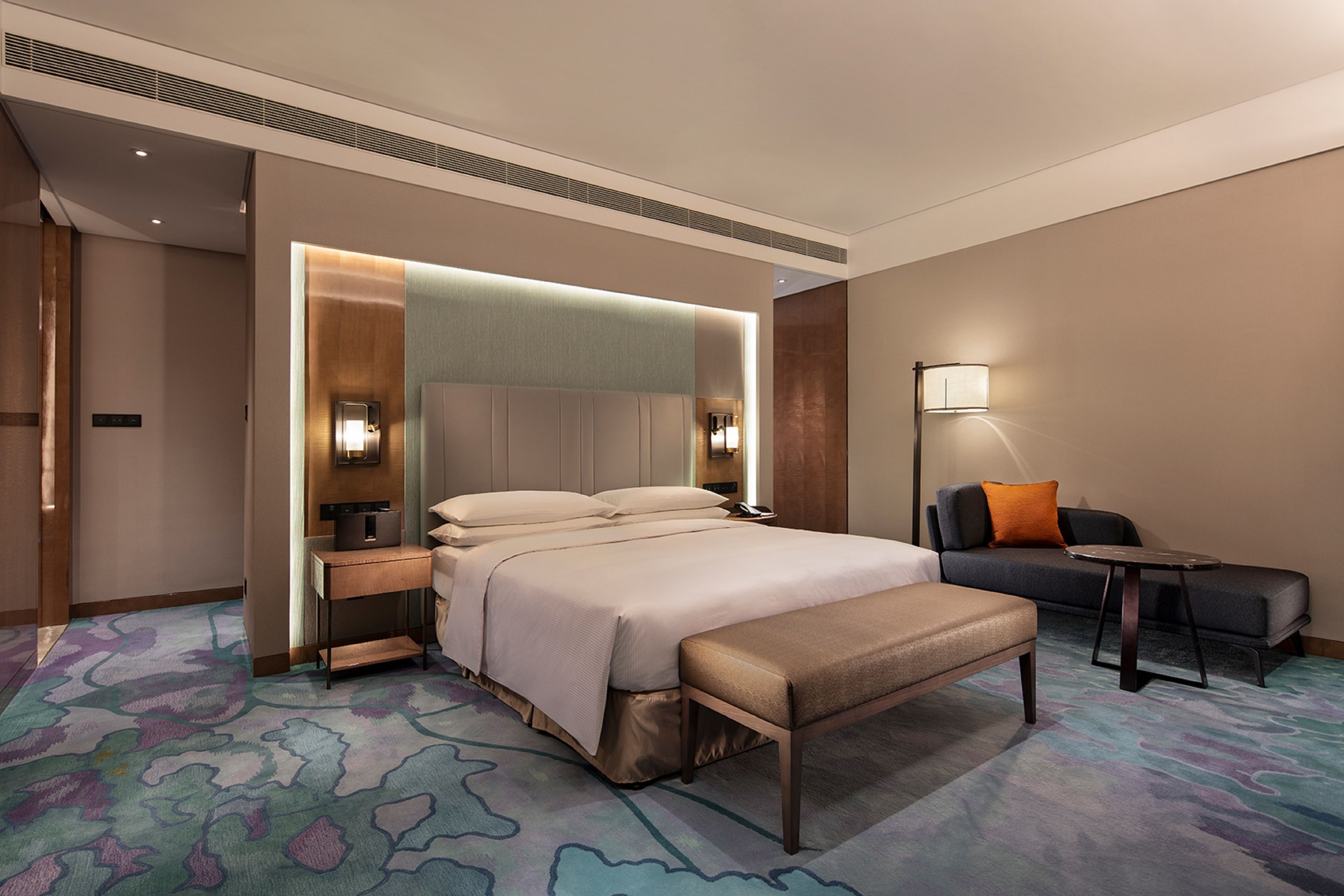 A modern hotel room with a large bed, bedside tables, a bench, a sofa, and a lamp.