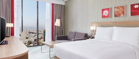 Habitación de hotel con una cama grande, un escritorio, una silla y vistas de la ciudad a través de amplias ventanas.