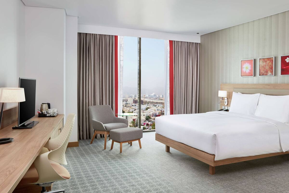 Ein Hotelzimmer mit einem großen Bett, einem Schreibtisch mit Computer, einem Sessel und Blick auf die Stadt durch große Fenster.