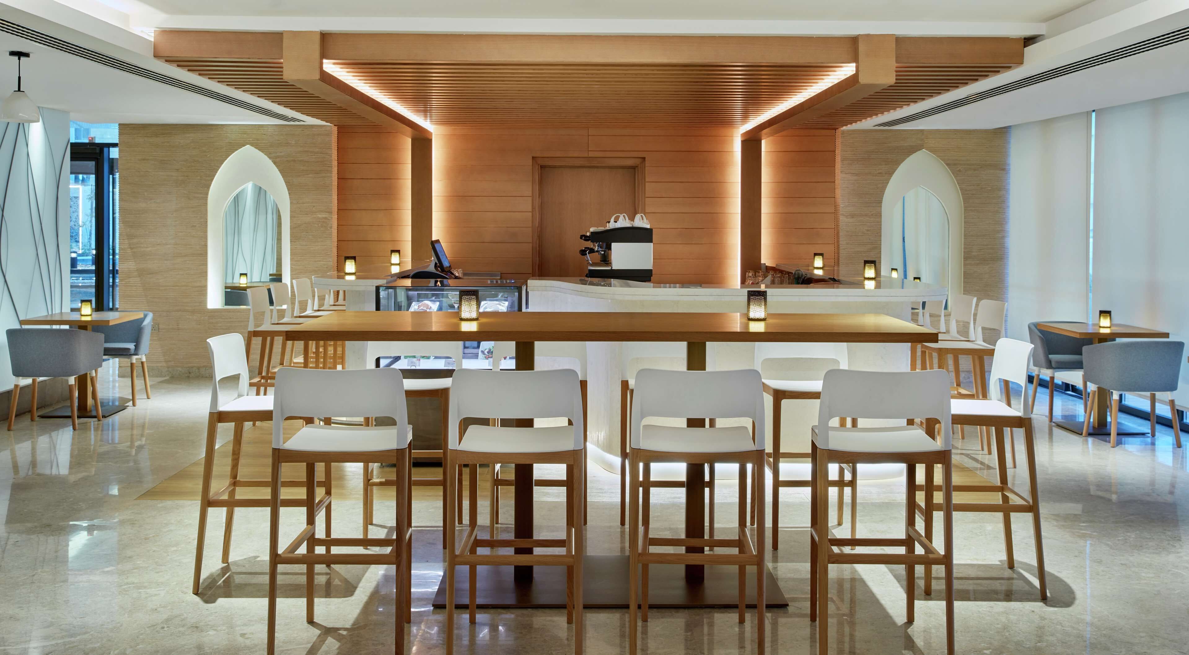 A modern bar area with a long wooden counter, white bar stools, and a central coffee machine.