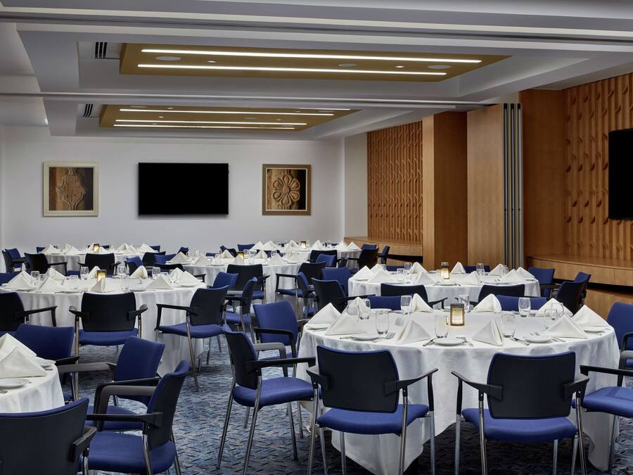 Un salón de conferencias con mesas redondas, sillas azules, manteles blancos y una pantalla grande en la pared.