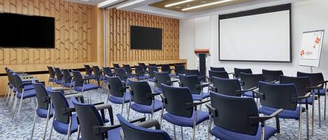 Un salón de conferencias con filas de sillas azules, una pantalla grande y un atril.