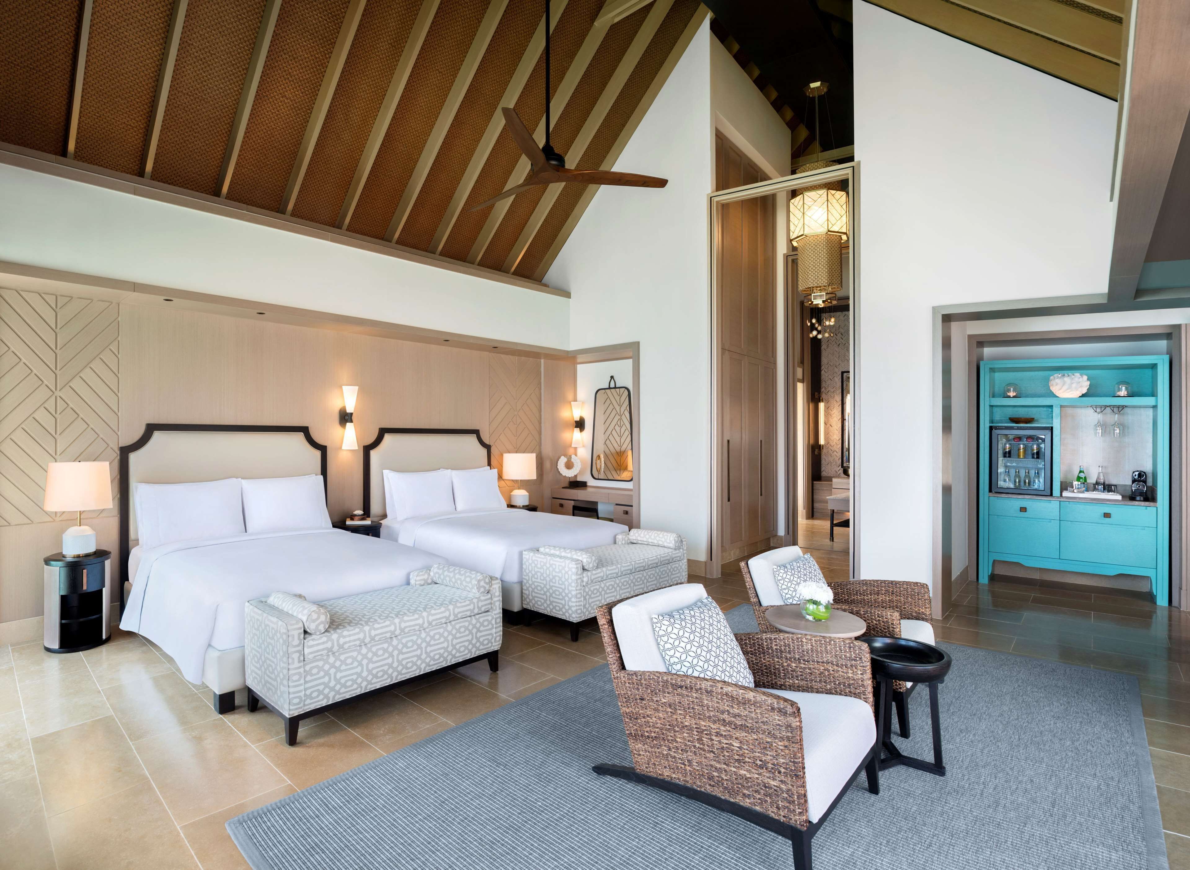 Habitación de hotel con dos camas, una zona de estar con muebles de mimbre, un minibar y un ventilador de techo.