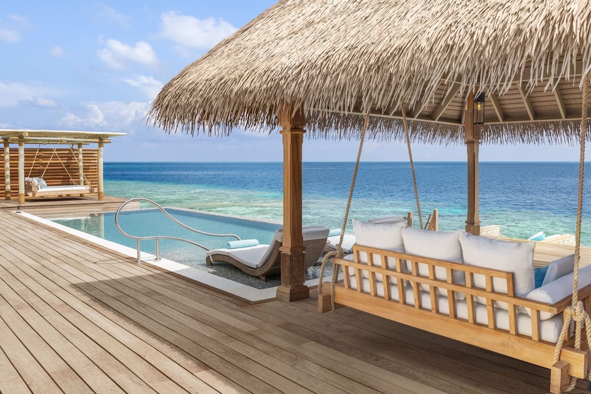 A wooden deck with a thatched-roof cabana, a swing, and lounge chairs overlooking a clear blue sea.