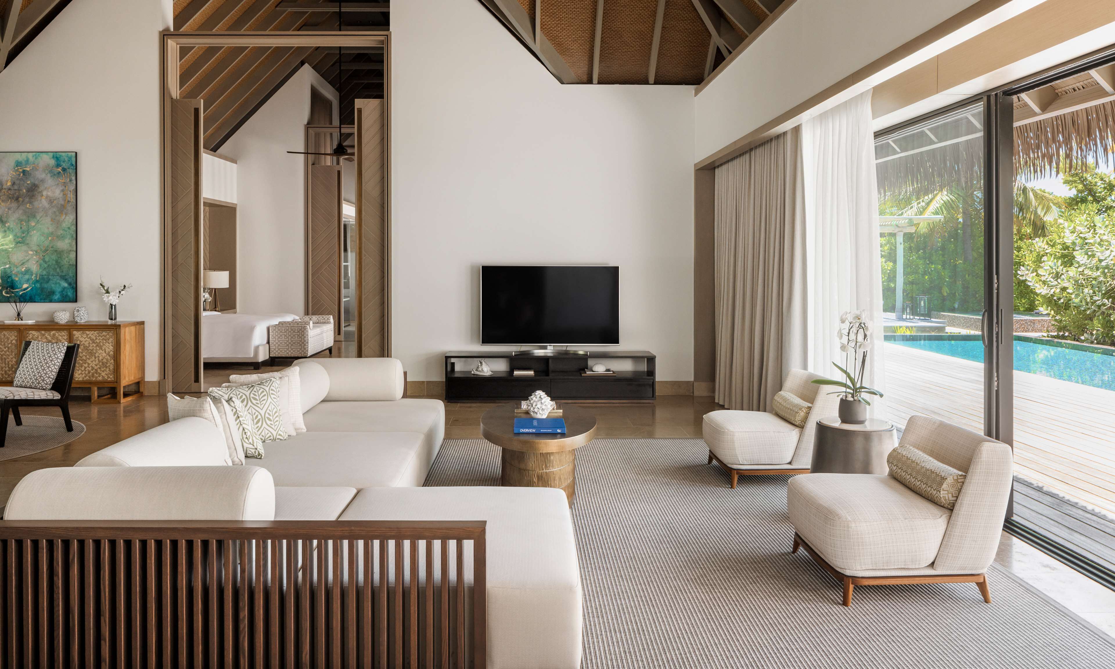 A modern living room with a large flat-screen TV, a sofa set, a coffee table, and a view of a swimming pool.