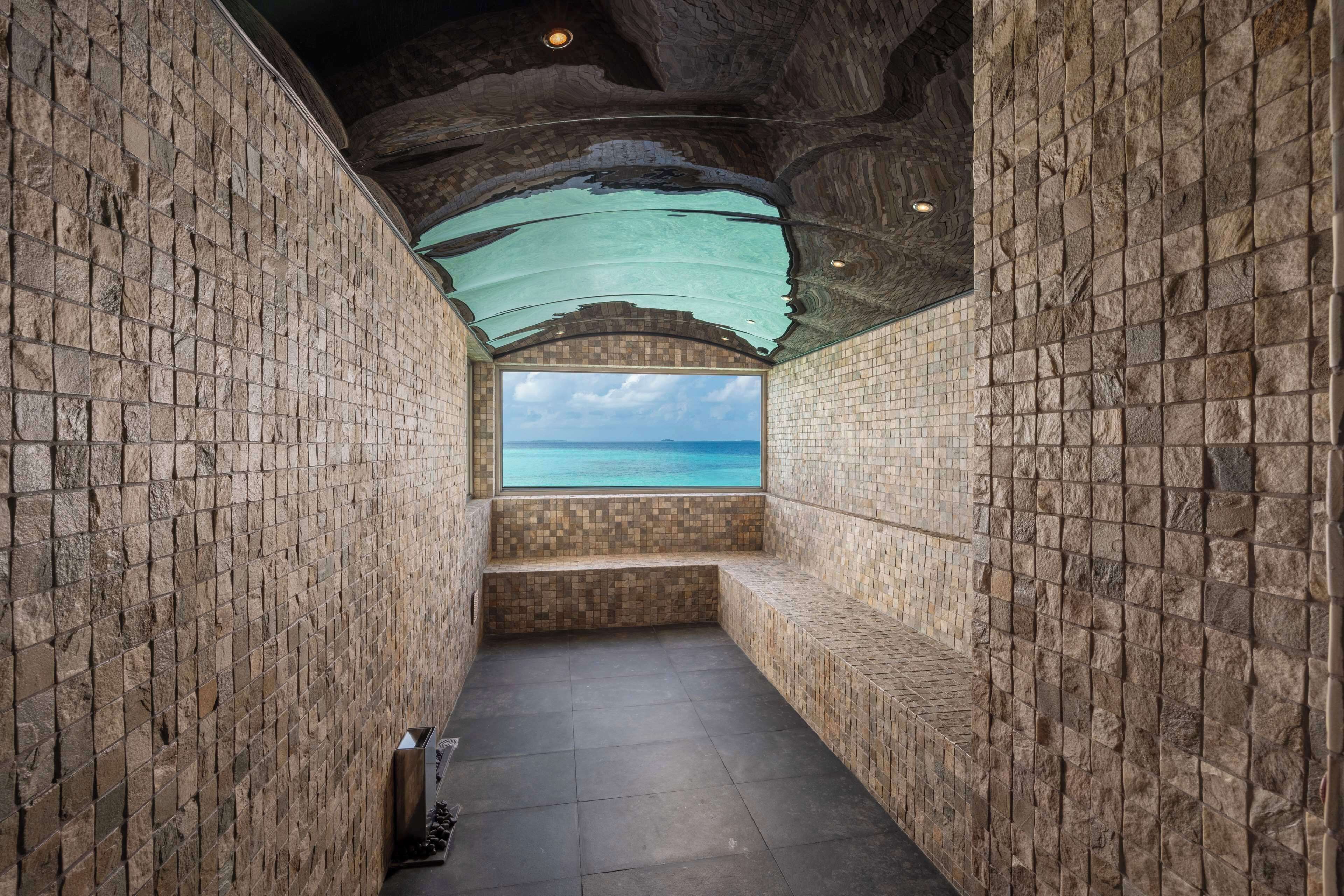 A narrow, stone-walled corridor with a bench and a view of the sea through a window.
