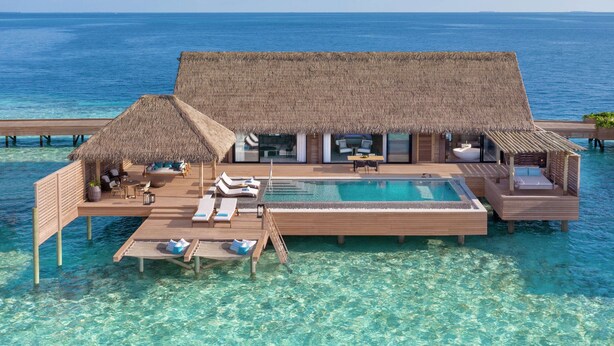 Una villa sobre el agua con piscina, sillones de descanso y estructuras de techo de paja.