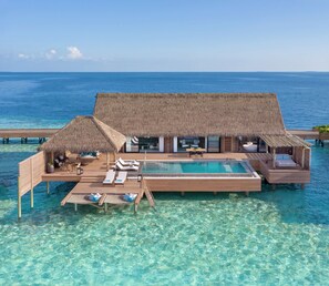 Una villa sobre el agua con piscina, sillones de descanso y estructuras de techo de paja.