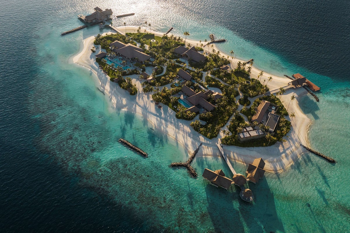 An aerial view of a resort island with clear turquoise waters, a sandy beach, and several resort buildings surrounded by palm trees.
