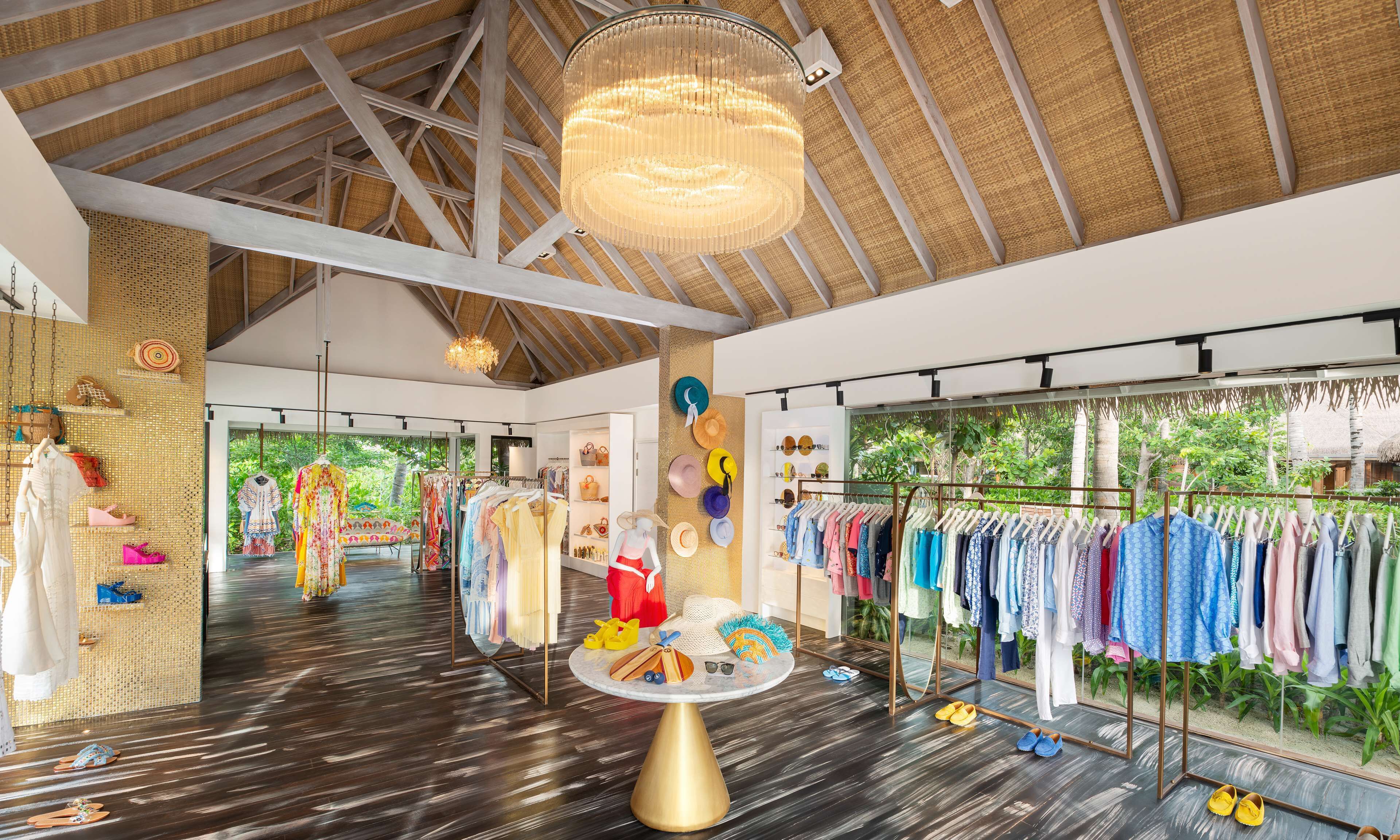 A boutique with a variety of clothing and accessories displayed on racks and tables, a large chandelier, and a view of greenery outside.