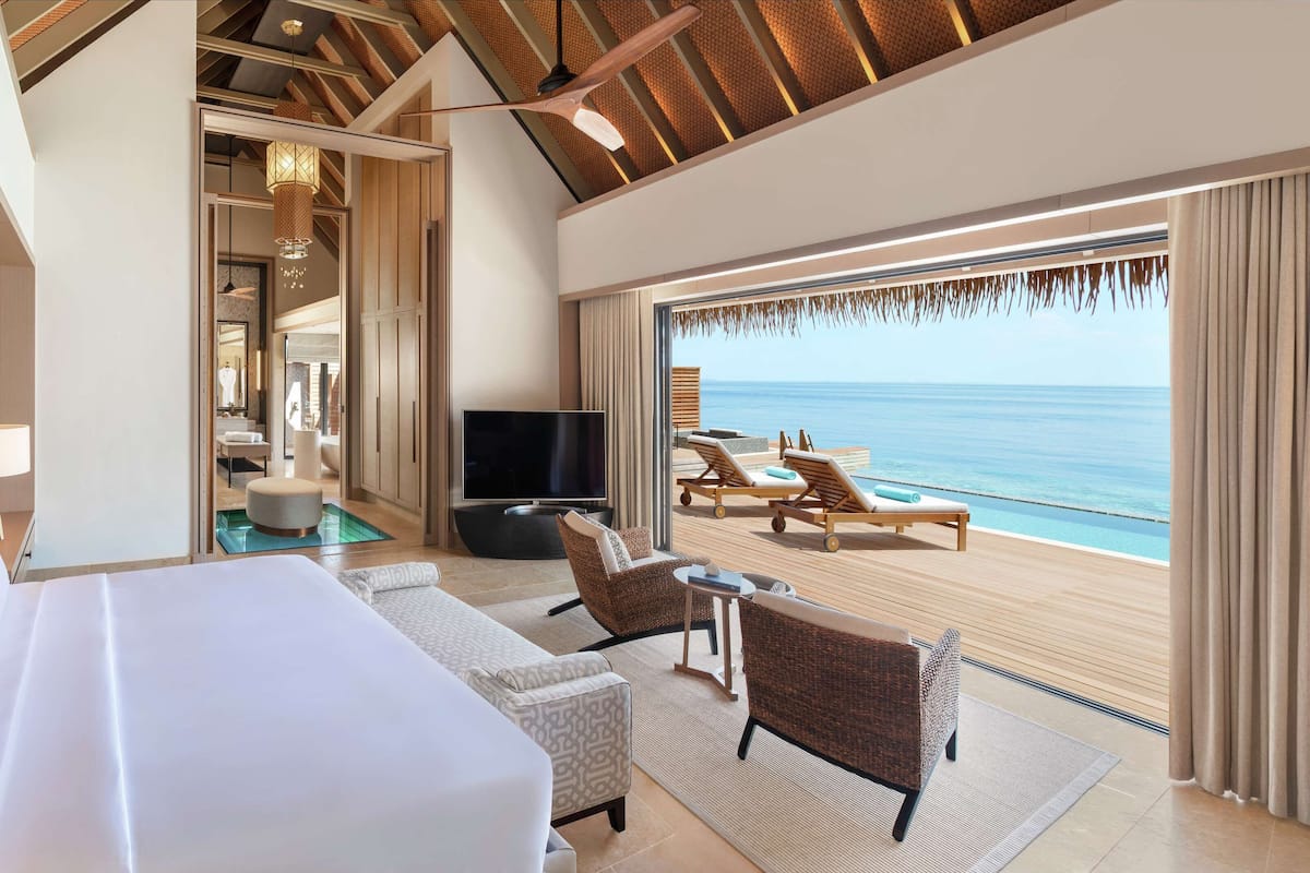 A hotel room with a large bed, a TV, a seating area with a view of the ocean, and a balcony with sun loungers.