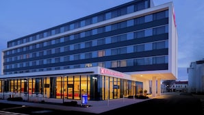 Un hotel moderno con fachada de vidrio, un área de restaurante visible con asientos rojos y una vista clara del logo Hilton Garden Inn.