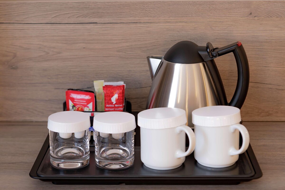 A tray with a stainless steel kettle, two instant coffee packets, and three white mugs on a wooden surface.