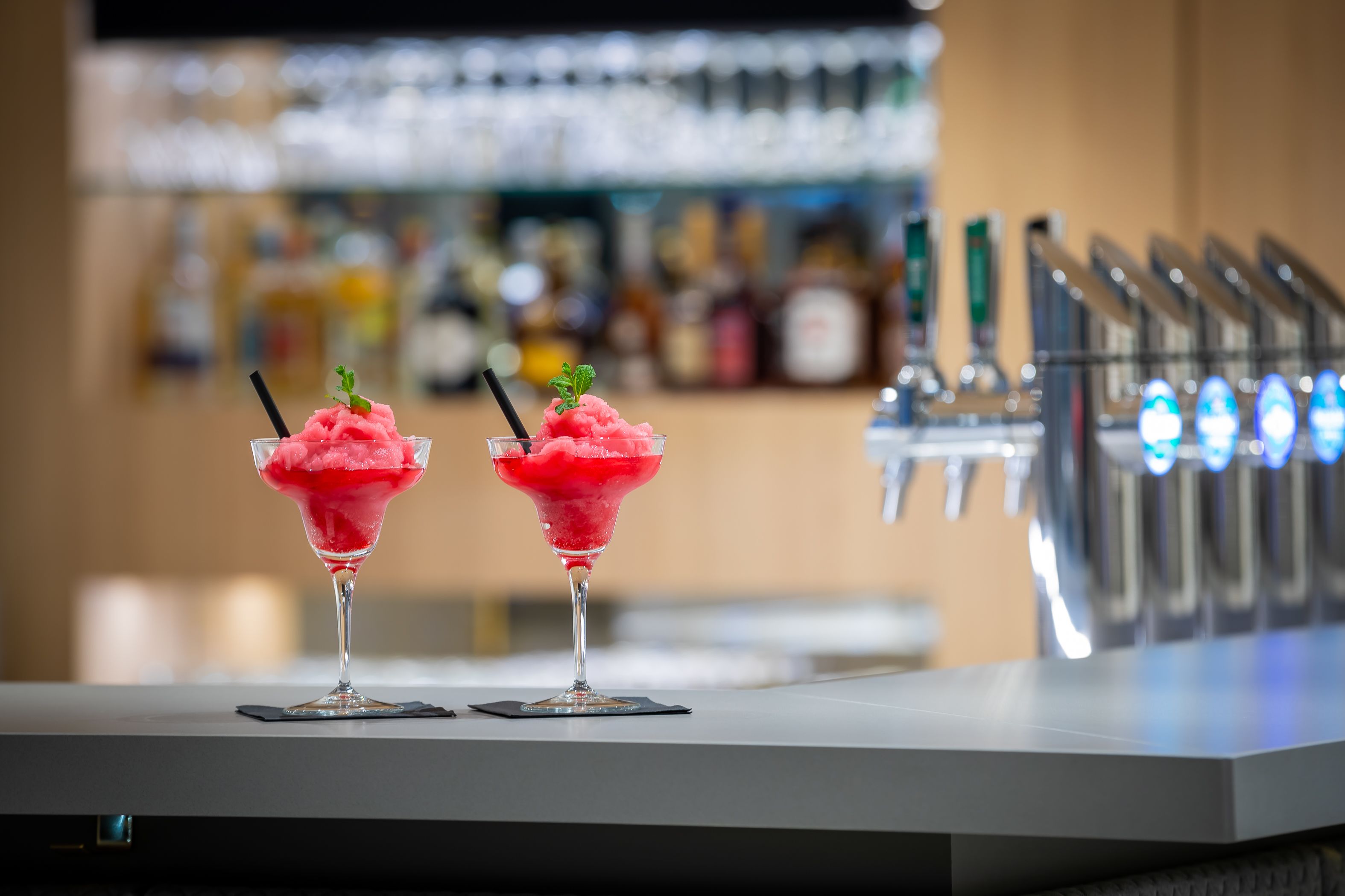 Two pink cocktails on a bar counter with a blurred background of bar equipment.