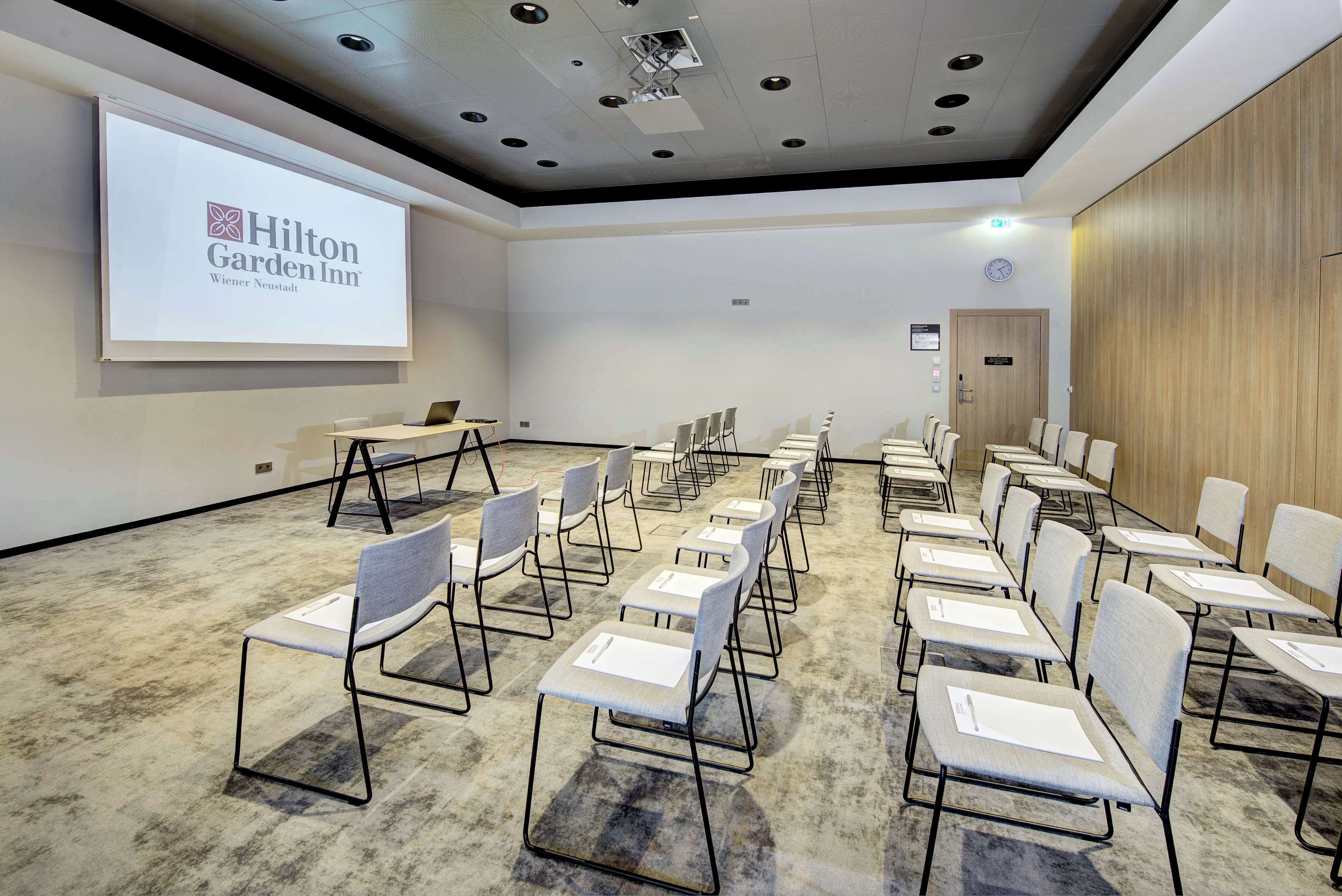 A conference room with rows of chairs, a projector screen displaying the Hilton Garden Inn logo, and a wooden panel wall.