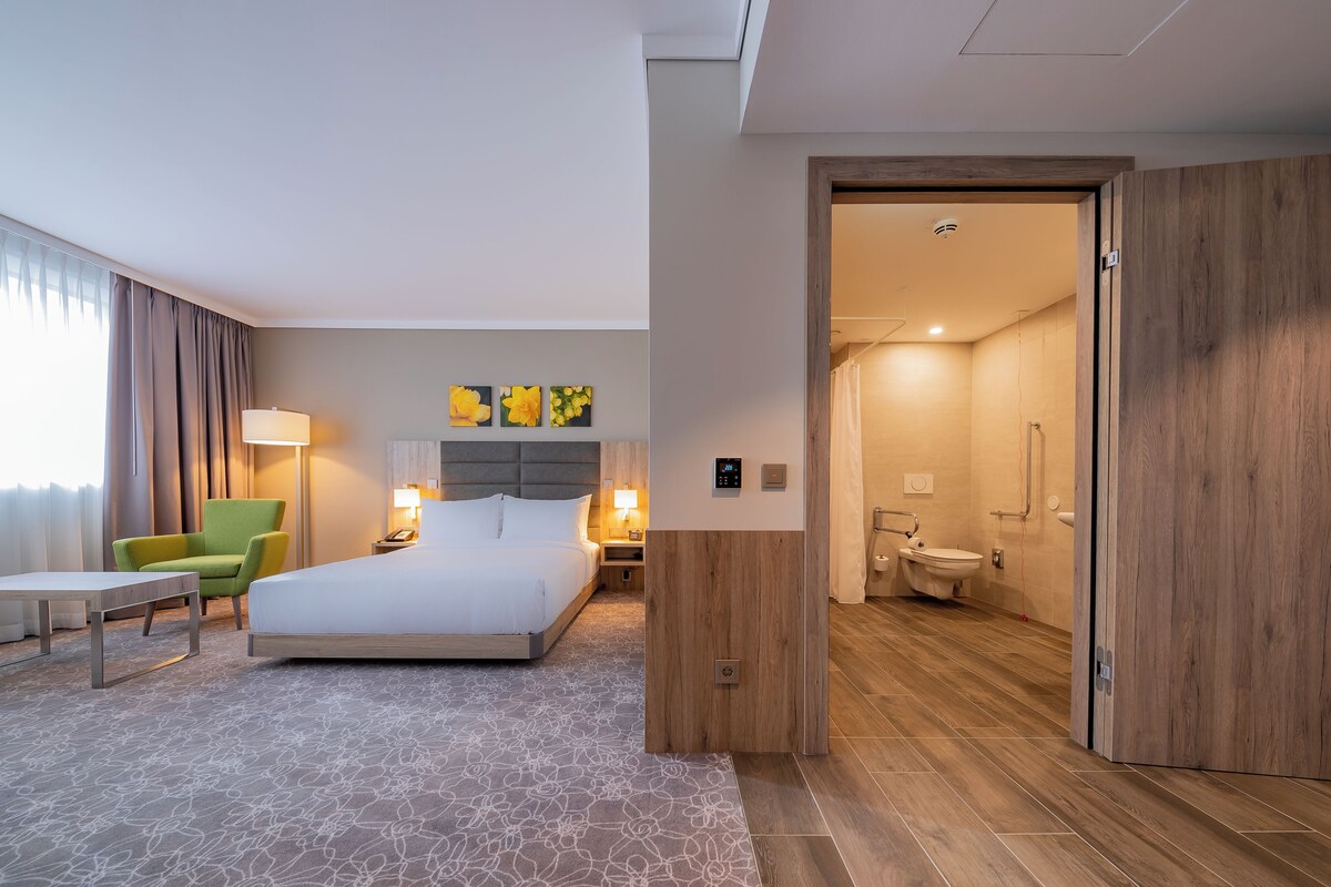 A hotel room with a large bed, a green armchair, a bedside table, and a lamp. There is a bathroom visible through an open door.