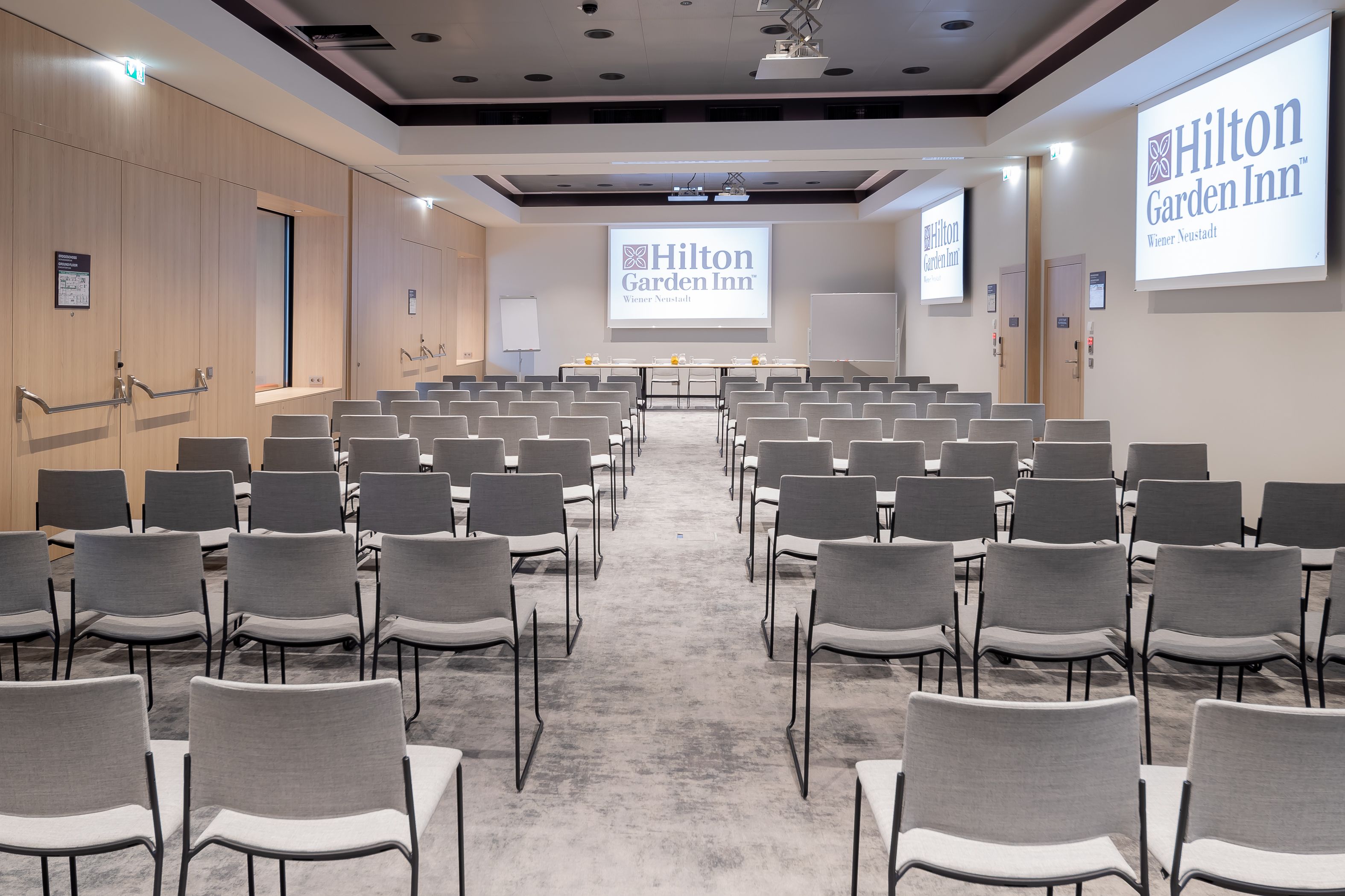 Une salle de conférence avec des rangées de chaises grises, un sol recouvert de moquette et un grand écran affichant le logo de l’hôtel Hilton Garden Inn.