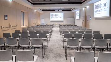 Une salle de conférence avec des rangées de chaises grises, un sol recouvert de moquette et un grand écran affichant le logo de l’hôtel Hilton Garden Inn.