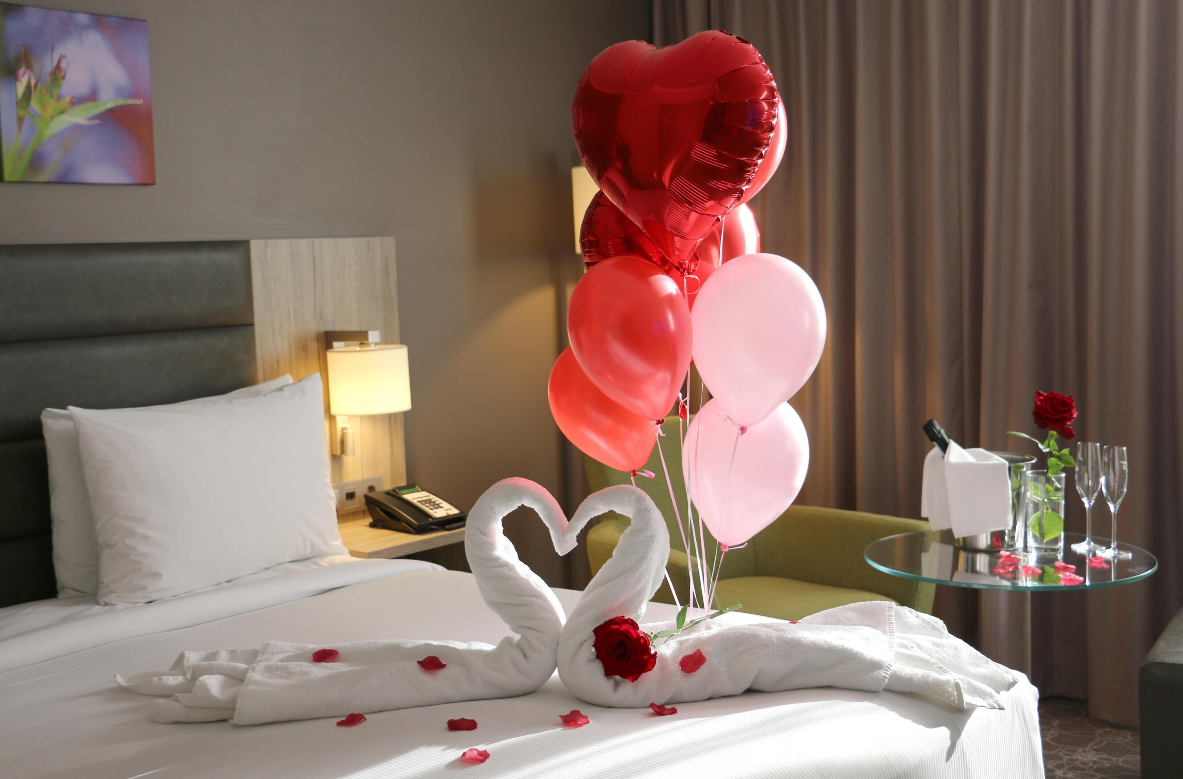 Habitación de hotel con una cama bien hecha, un ramo de globos rojos y rosas, una decoración de toallas en forma de cisne con una rosa, y una pequeña mesa con un florero de rosas rojas y copas de champán.