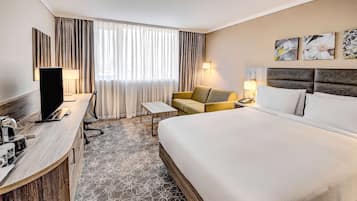 Habitación de hotel con una cama grande, un escritorio con computadora, una silla, una mesita y un ventanal con cortinas.