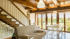 Een gezellige woonkamer met een houten trap, een bank, een eettafel met stoelen en grote ramen met uitzicht op het groen.