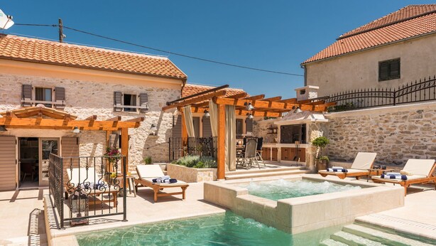 Un espace piscine agrémenté de chaises longues, d’une pergola et de bâtiments en pierre surmontés de toits en tuiles.