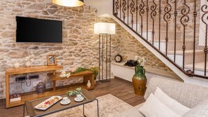 Un salon avec un mur en pierre, un téléviseur à écran plat, une console en bois sur laquelle est posé un cadre photo, une lampe sur pied, un escalier avec des rampes en fer forgé et un canapé.