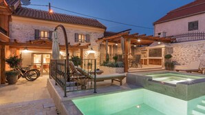 Une cour avec une piscine, des espaces de détente en plein air et un bâtiment en pierre surmonté d’un toit en tuiles.