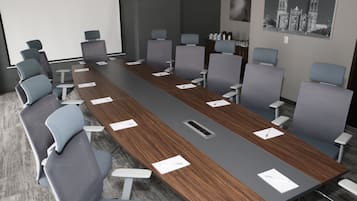 Un salón de conferencias con una larga mesa, sillas grises, una pantalla de proyector y un cuadro en la pared.