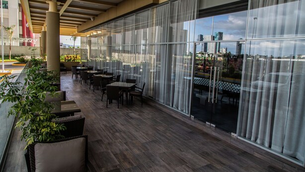 An outdoor seating area with tables and chairs, large windows, and potted plants. - Hampton Inn & Suites by Hilton Puebla (San Andrés Cholula)