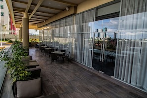 An outdoor seating area with tables and chairs, large windows, and potted plants. - Hampton Inn & Suites by Hilton Puebla (San Andrés Cholula)