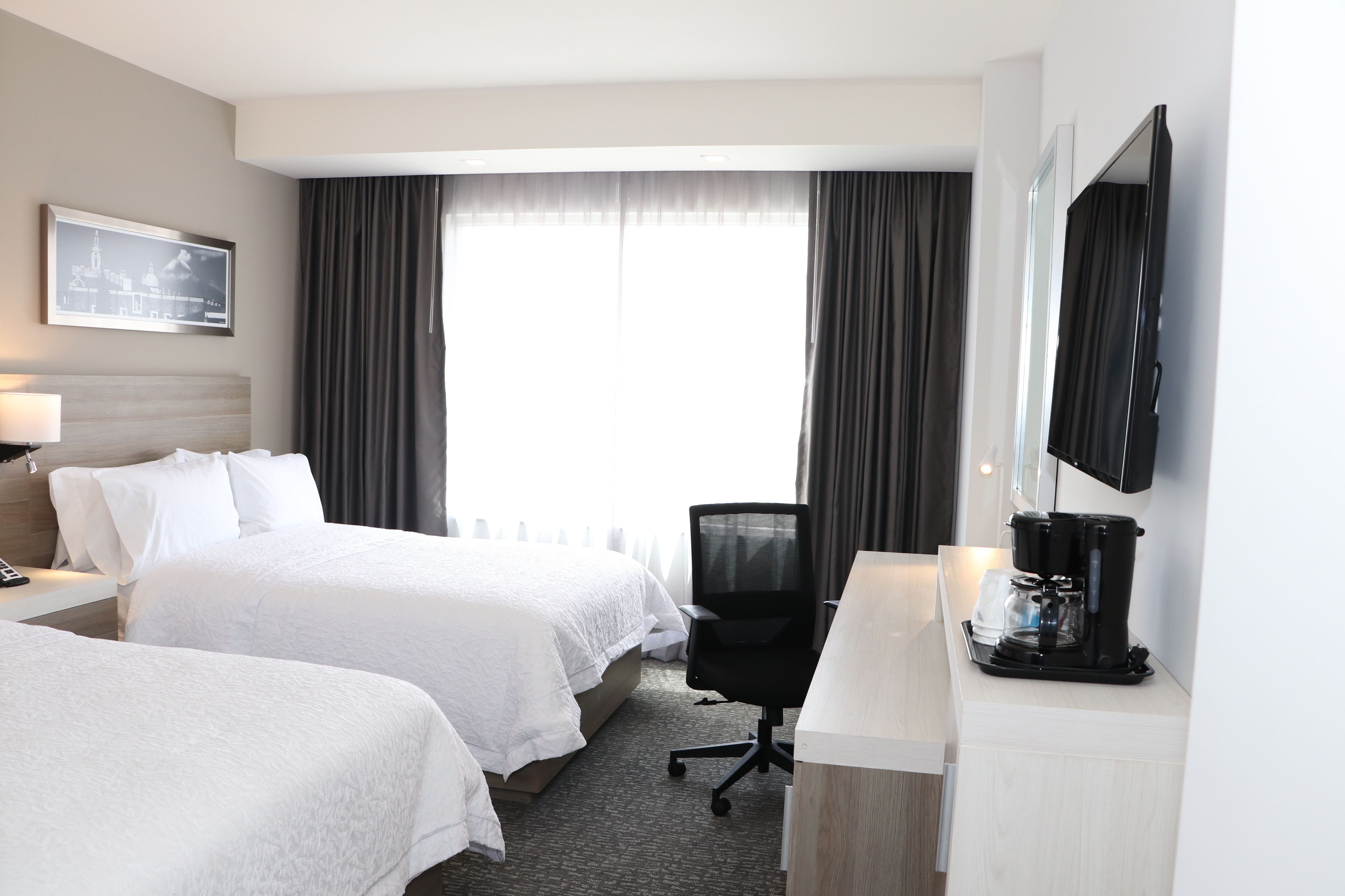 A hotel room with two beds, a desk with a coffee maker, a TV, and a window with curtains.