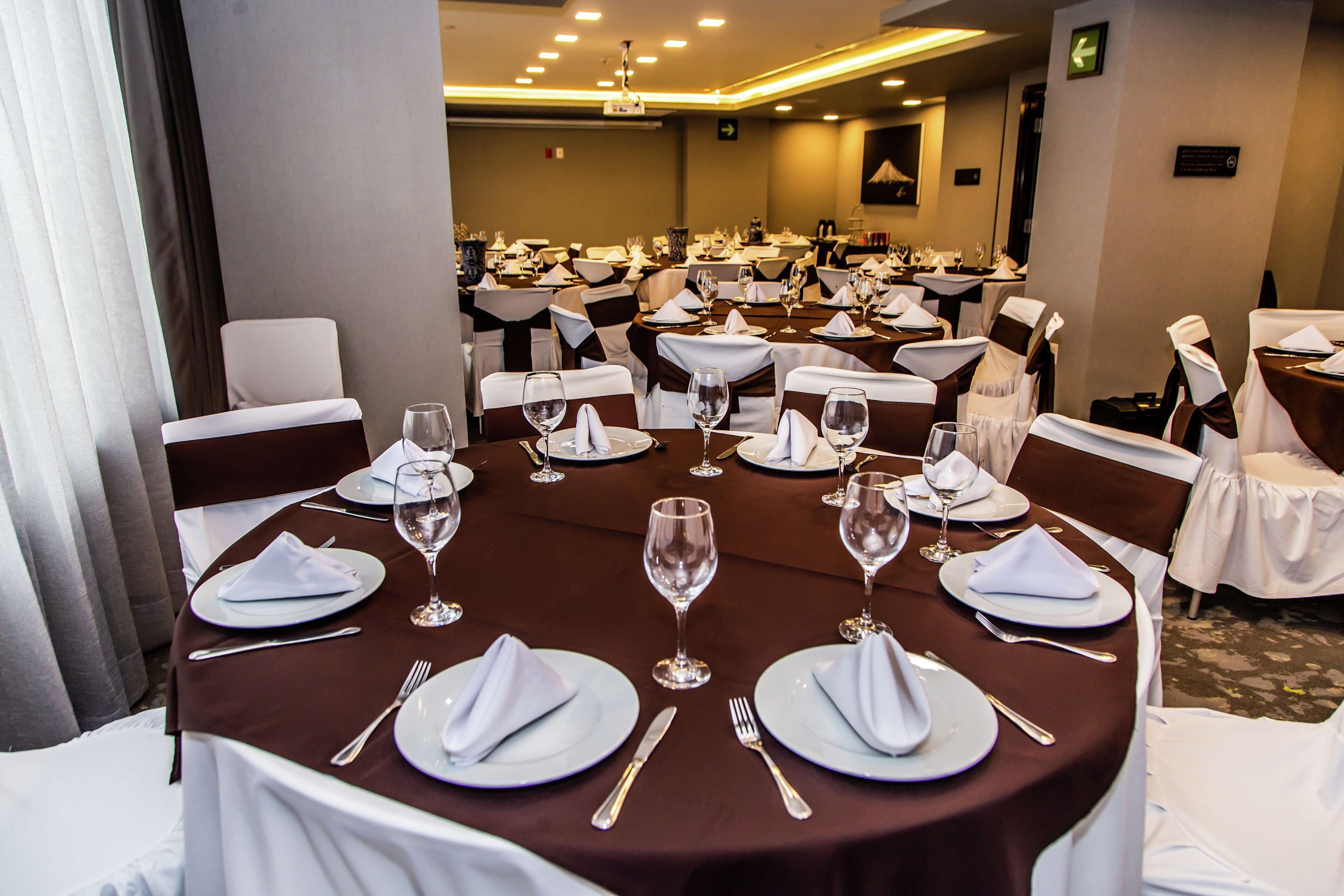 Un salón de banquetes con mesas redondas dispuestas para un evento formal, con manteles blancos, servilletas y copas de vino.